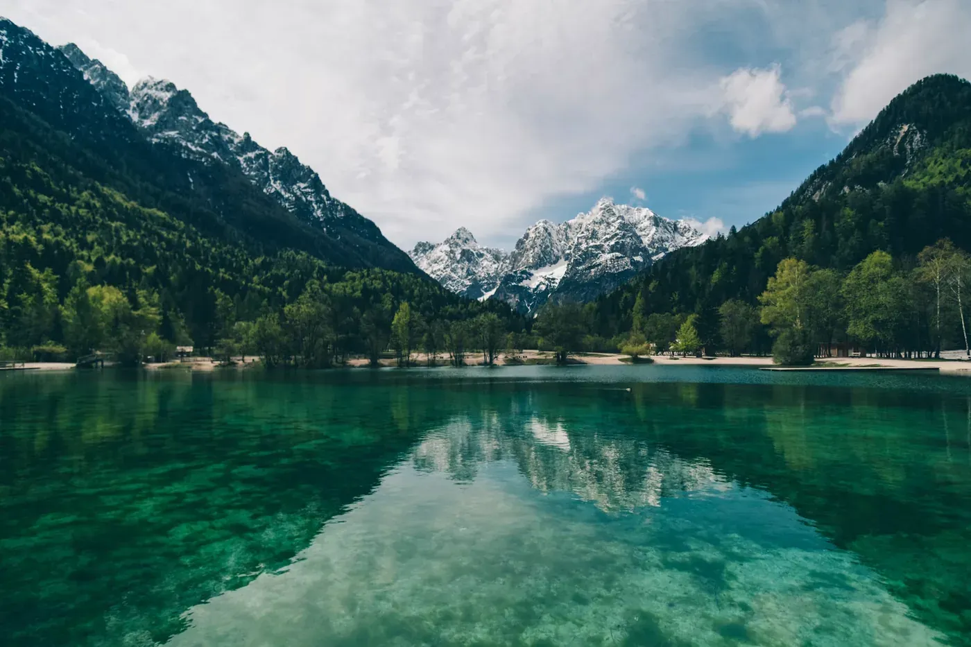 A turquoise alpine lake with snow-capped mountains reflected in still water, framed by forested slopes.
