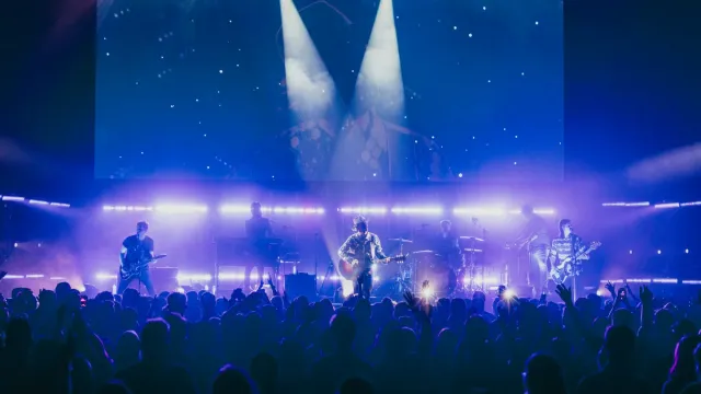 Volle zaal voor een popconcert met blauw en paars podiumlicht; de band staat groot op het LED-scherm op de achtergrond.