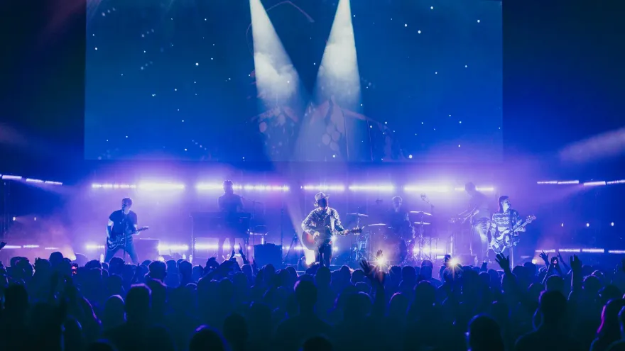 Volle zaal voor een popconcert met blauw en paars podiumlicht; de band staat groot op het LED-scherm op de achtergrond.