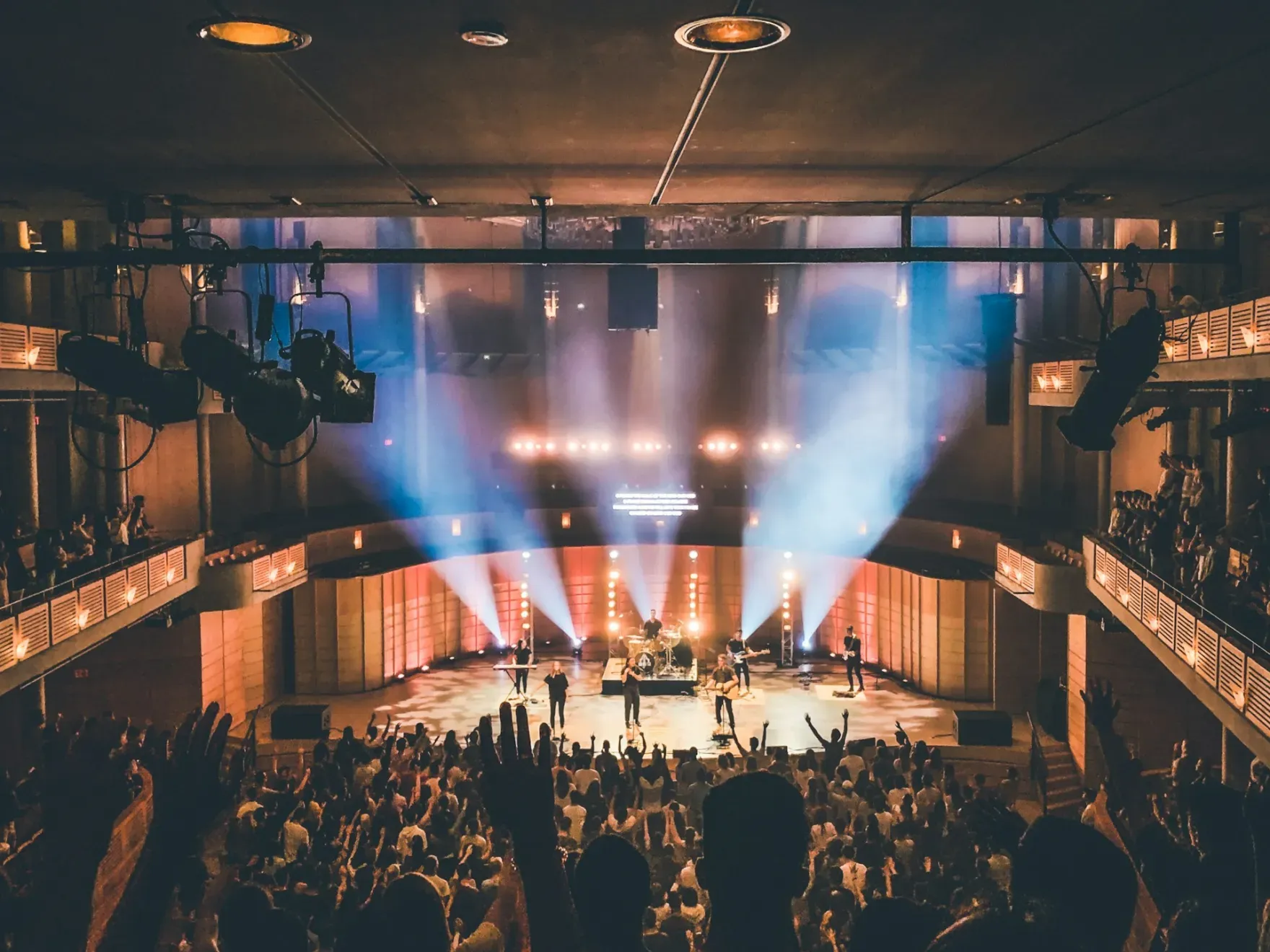 Middelgrote concertzaal van bovenaf gezien: een band op het podium, het publiek met de armen omhoog en warme, blauwe en oranje lichtbundels in de zaal.