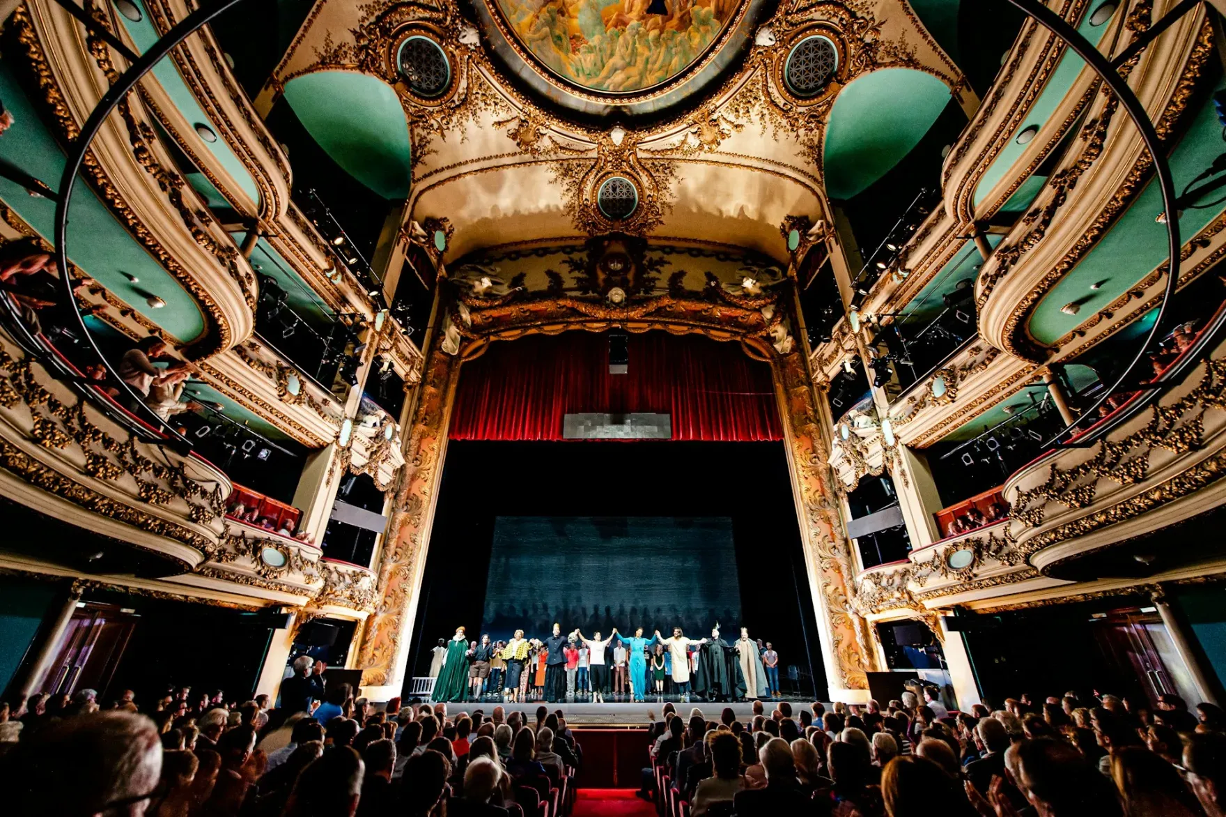 Rijk gedecoreerd klassiek theater met gouden ornamenten en groen interieur; het ensemble maakt na de voorstelling een buiging op het podium voor een volle zaal.